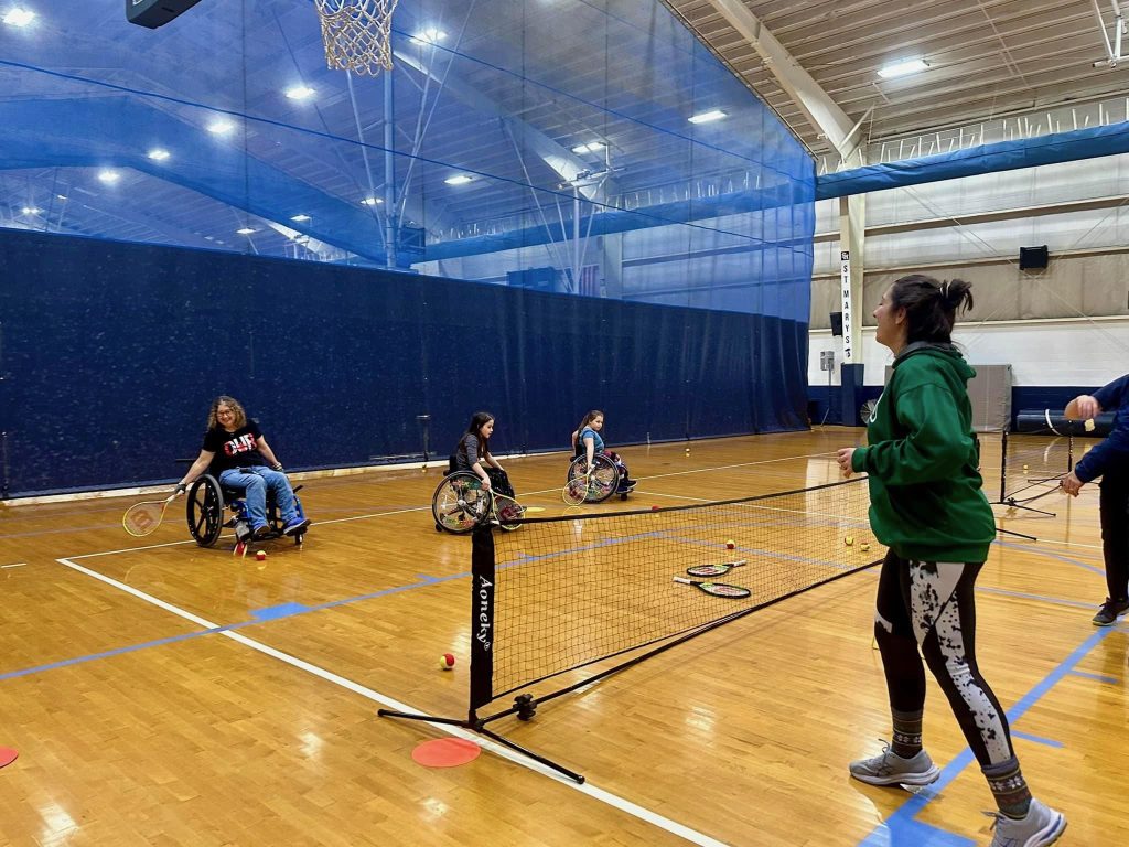 GBAS volunteers tosses a a ball to youth and adult wheelchair tennis players in a gym.