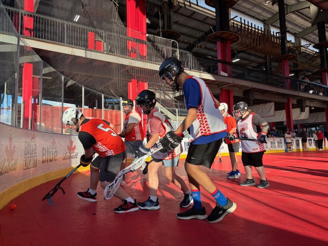 Stand-up lacrosse players competing for the ball in the corner of a rink.